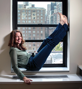 Robin Weigert sits on the inside sill of a window that looks out on a urban view, with her bare feet propped up against the right side of the window